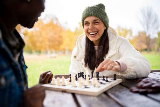 Smiling woman plays chess board outdoors with friend, autumn background.