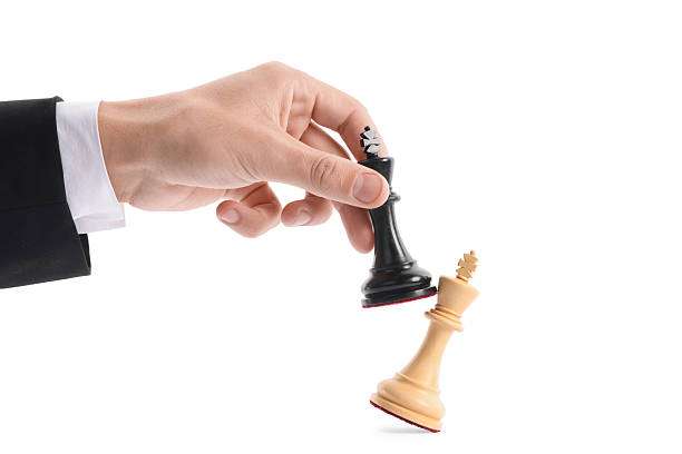 Hand holding a black king chess piece, triumphing over a white king on a chess board.